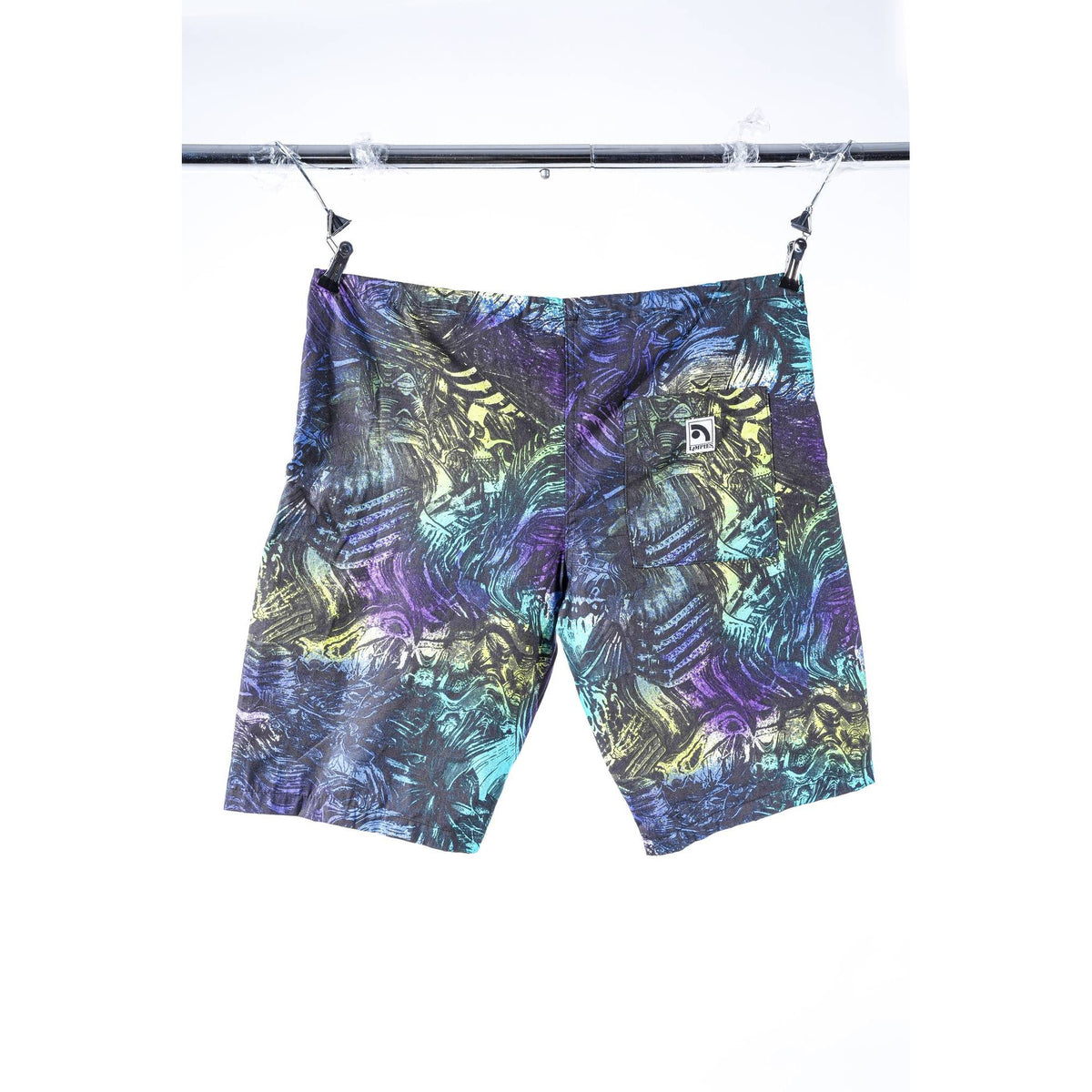 A pair of Limpies baggy skate shorts with an abstract, multicolor pattern of purple, teal, yellow, and black, hanging from a black metal clothesline against a white background. A small white square Limpies logo patch is visible on the upper right thigh.