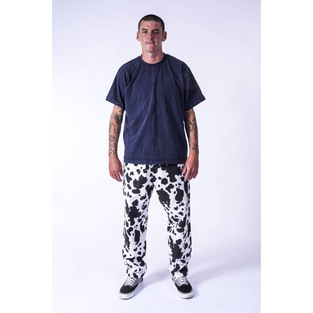 A street skater man modeling a dark navy blue t-shirt and black and white cow-print Limpies baggy skate pants against a plain white studio background.