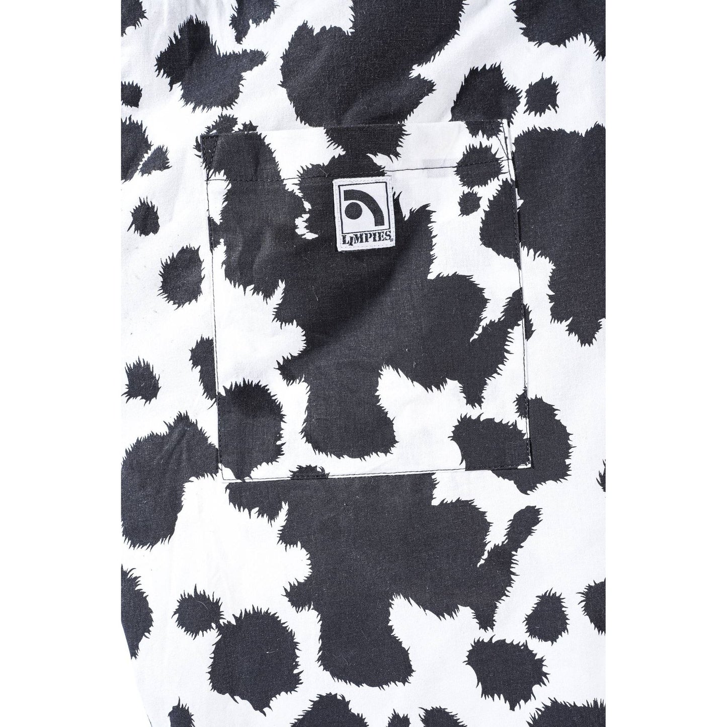 Close-up of a black and white cow-print Limpies baggy skate short fabric garment, showing a front pocket with a square Limpies logo patch.