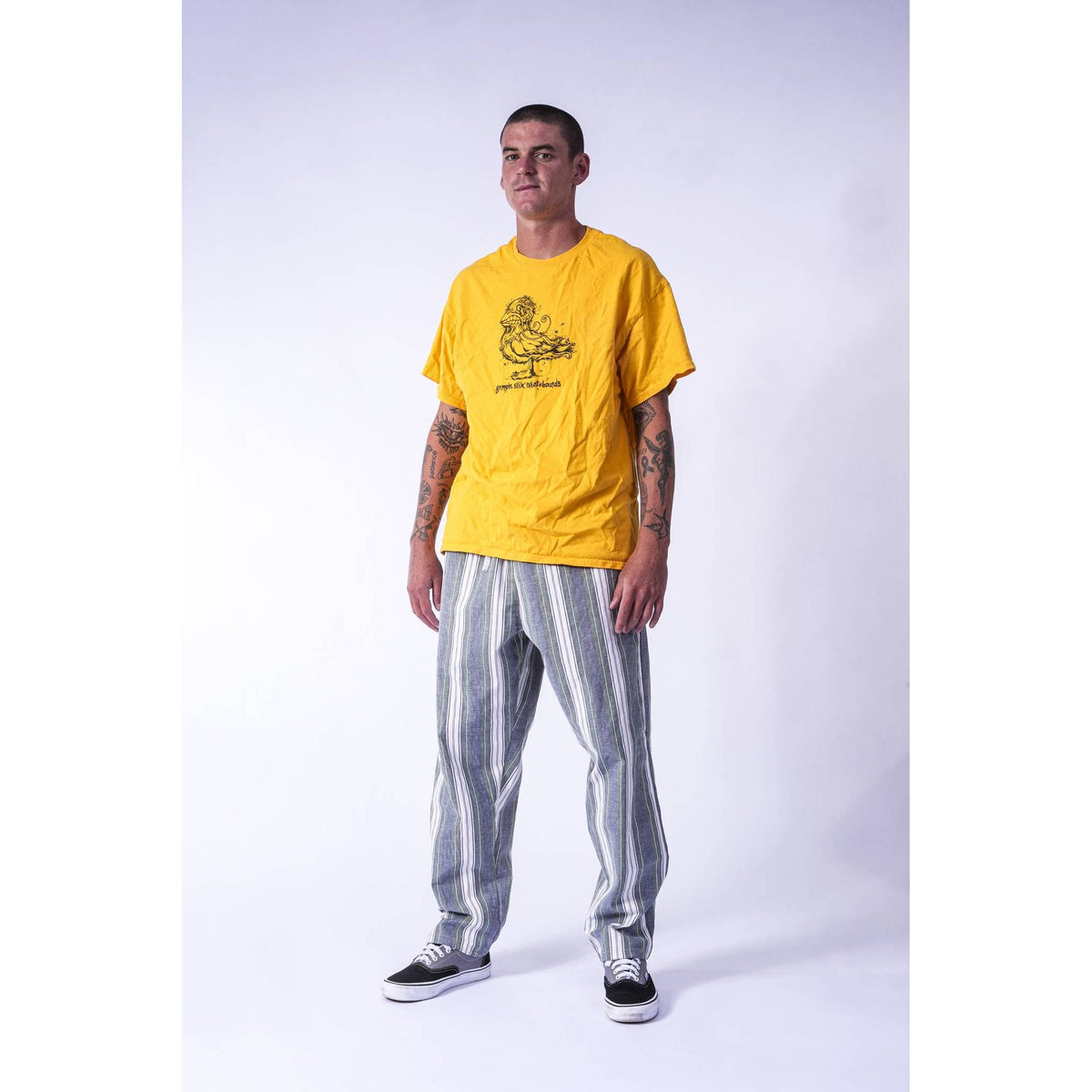 A man modeling a bright yellow t-shirt with a black graphic print on the chest, paired with gray and white vertically striped pants and black sneakers, standing against a light gray studio background.