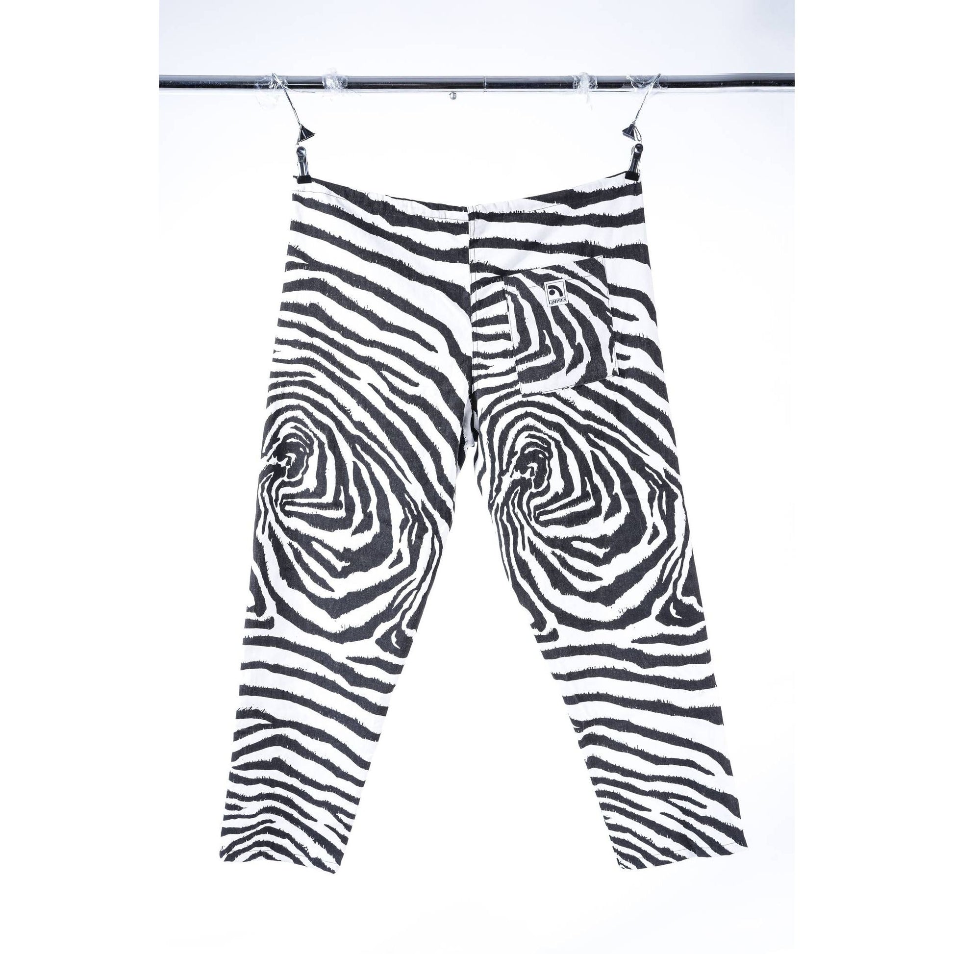 Black and white zebra-print cropped baggy skate pants hanging from clips on a black rail against a white background.