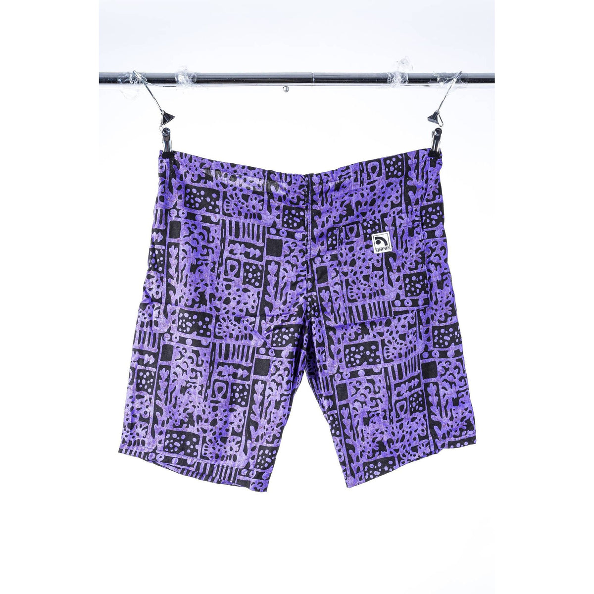 Purple and black patterned Limpies baggy skate shorts hanging from clips on a metal hanger bar against a white background.