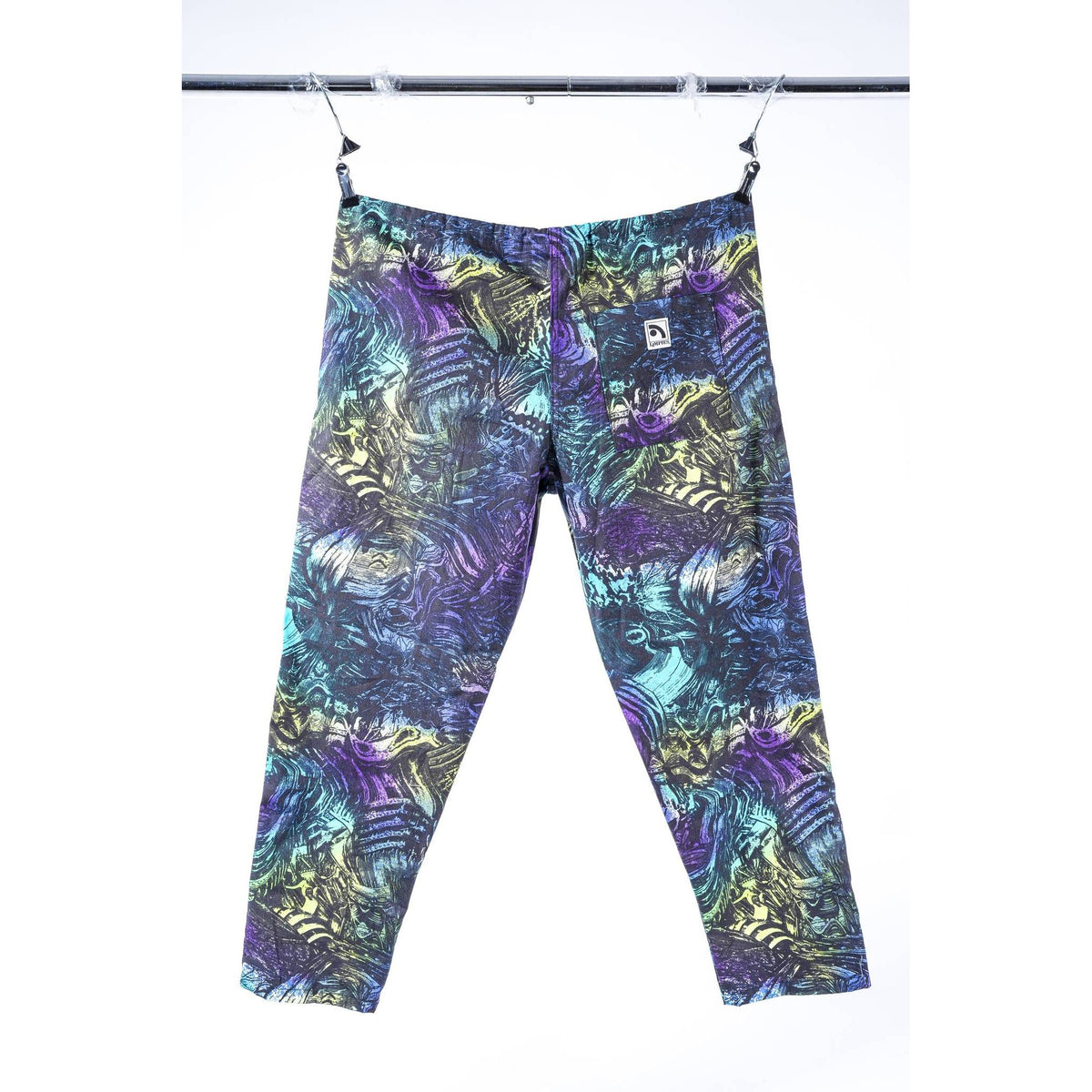A pair of  Limpies baggy skate pants with an abstract, multicolored pattern featuring purple, teal, green, and yellow, hanging from clips on a metal rack against a white background.