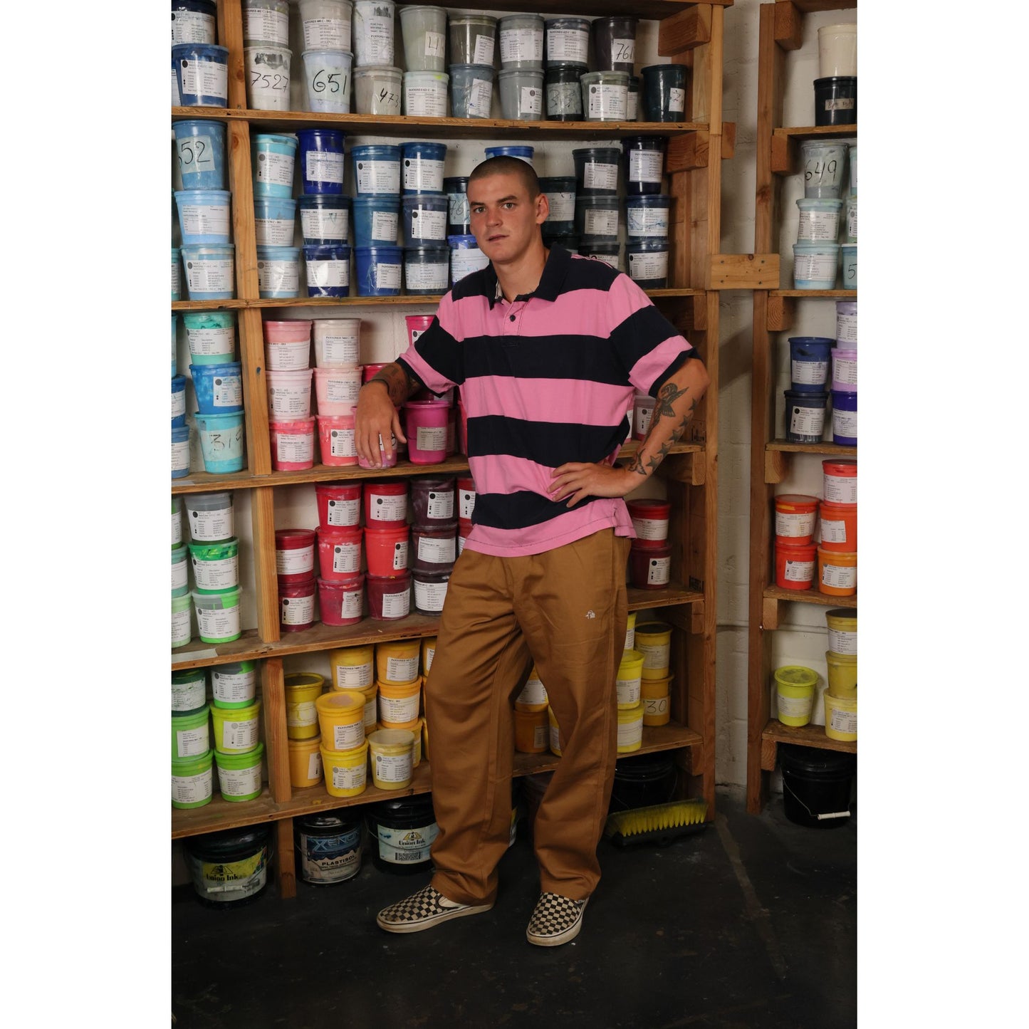 Man wearing baggy skate pants standing in front of shelves filled with various containers in a storage room.