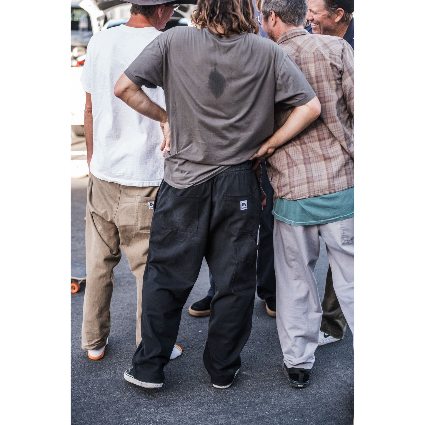 Man in a gray shirt and black baggy skate pants standing with his back to the camera, surrounded by other people on a street.