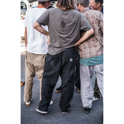 Man in a gray shirt and black baggy skate pants standing with his back to the camera, surrounded by other people on a street.