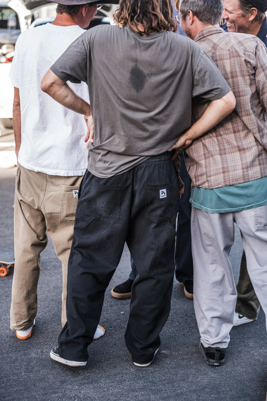 Man in a gray shirt and black baggy skate pants standing with his back to the camera, surrounded by other people on a street.