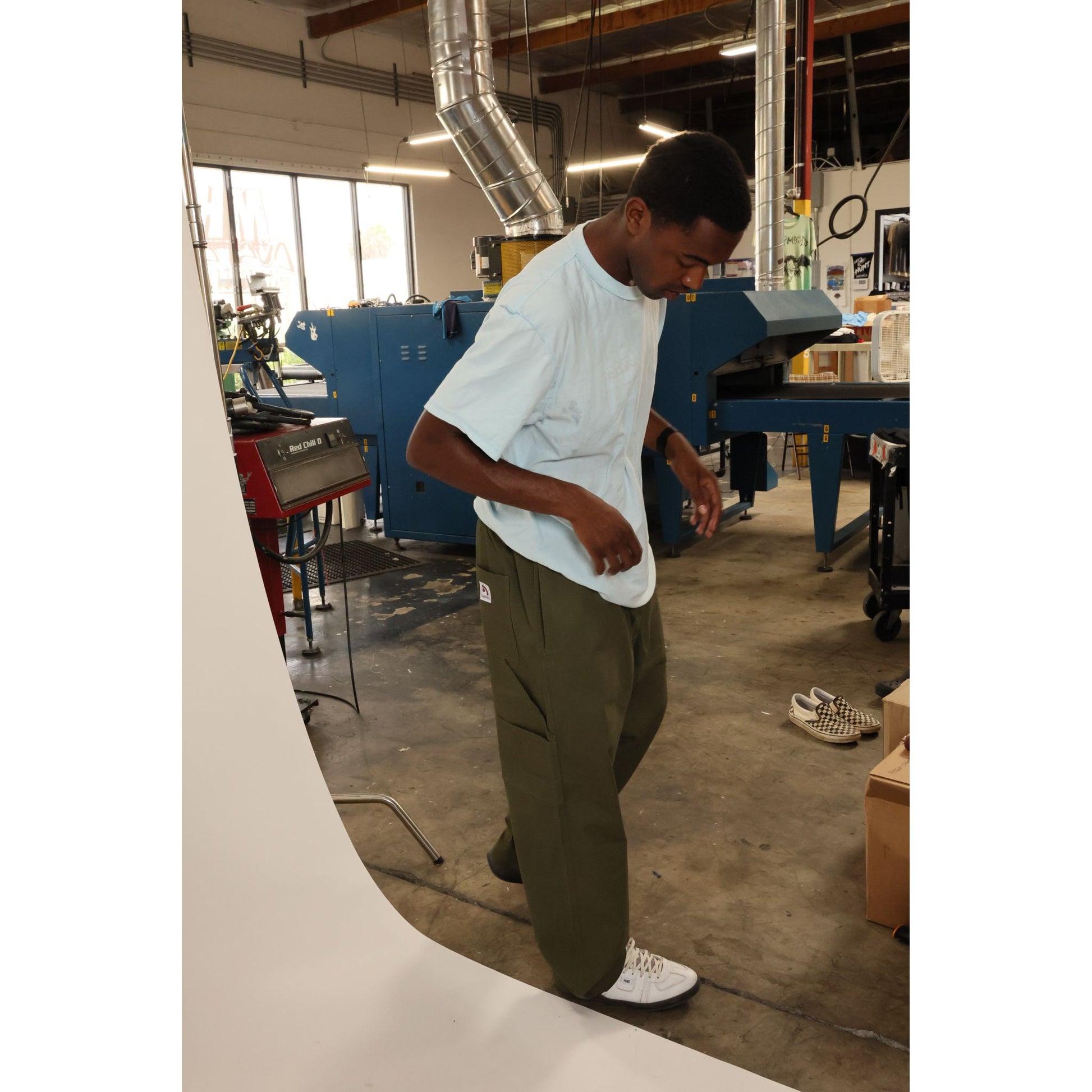 Person wearing olive green baggy skate pants standing in a workshop with machinery and tools in the background