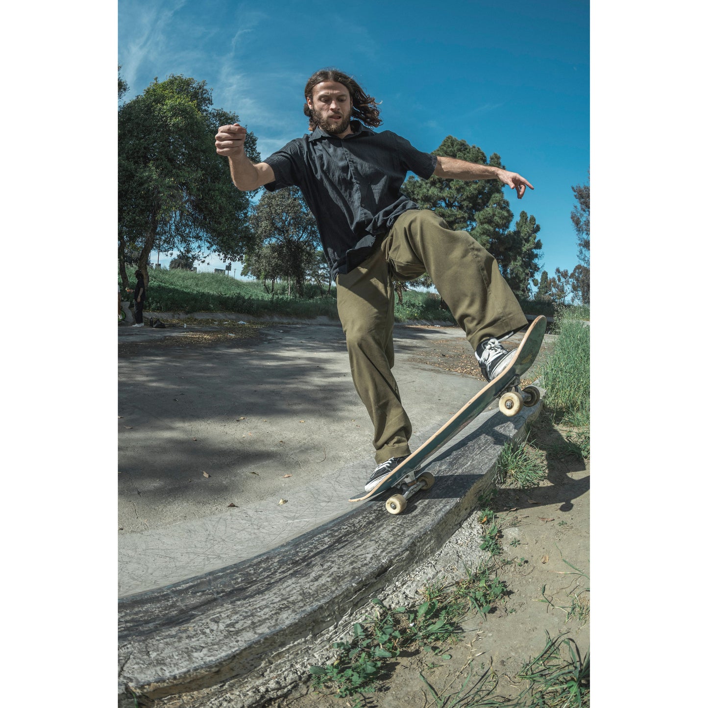 Person skateboarding in baggy skate pants on a concrete ledge with trees and blue sky in the background