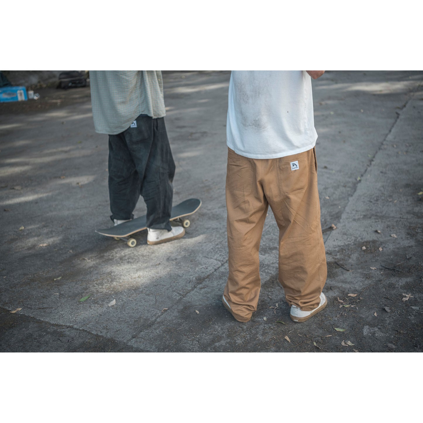 Two people wearing baggy skate pants on a street with one person on a skateboard