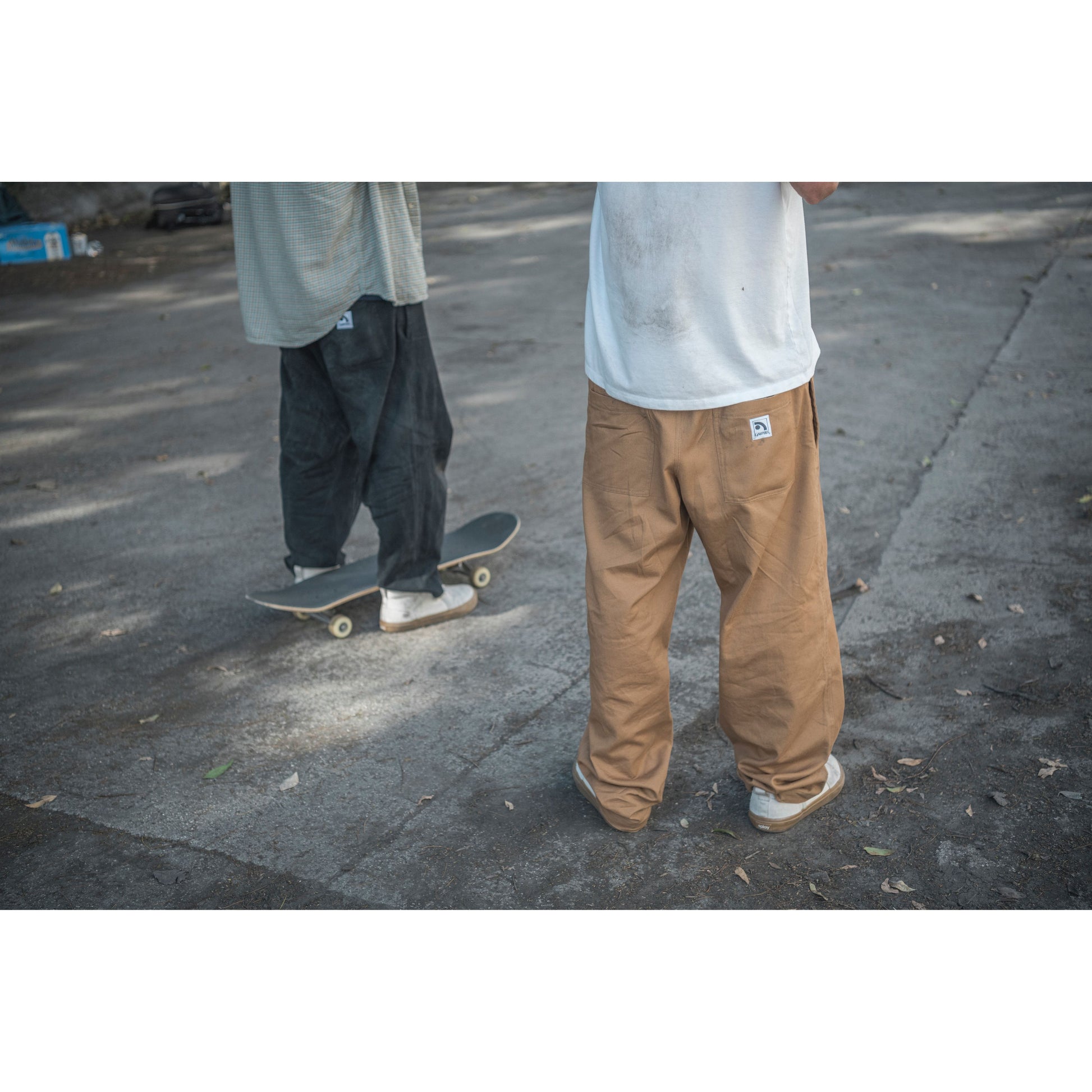 Two people wearing baggy skate pants on a street with one person on a skateboard