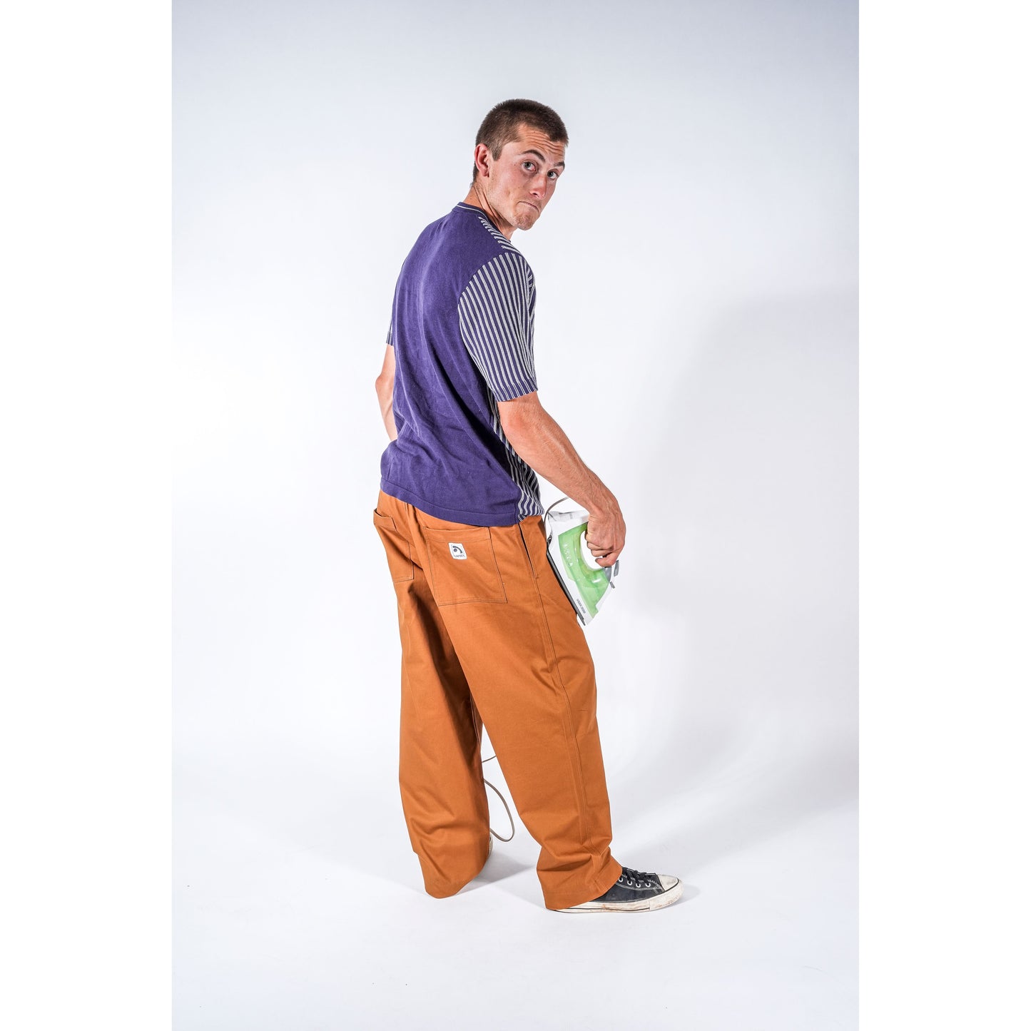 A man wearing a purple and gray striped t-shirt, beige wide-leg baggy skate pants, and black sneakers, holding a green and white iron in his pocket, photographed against a white studio background.