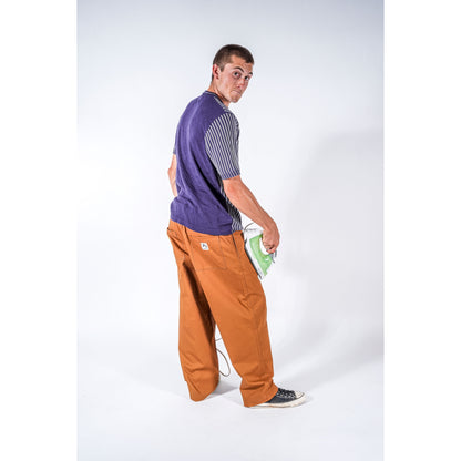 A man wearing a purple and gray striped t-shirt, beige wide-leg baggy skate pants, and black sneakers, holding a green and white iron in his pocket, photographed against a white studio background.