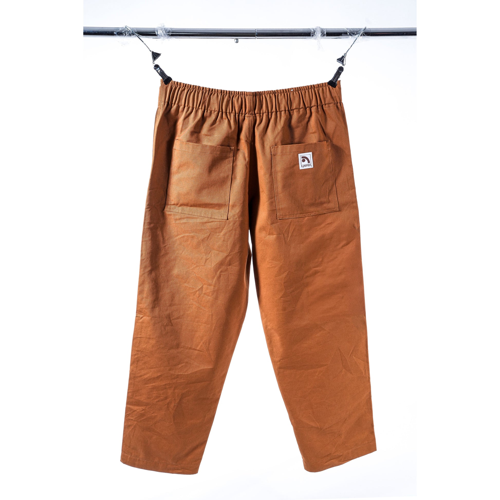 A pair of brown elastic-waist baggy skate pants with front patch pockets, hanging from a black metal bar against a white background. A small white brand label is visible on the right pocket.