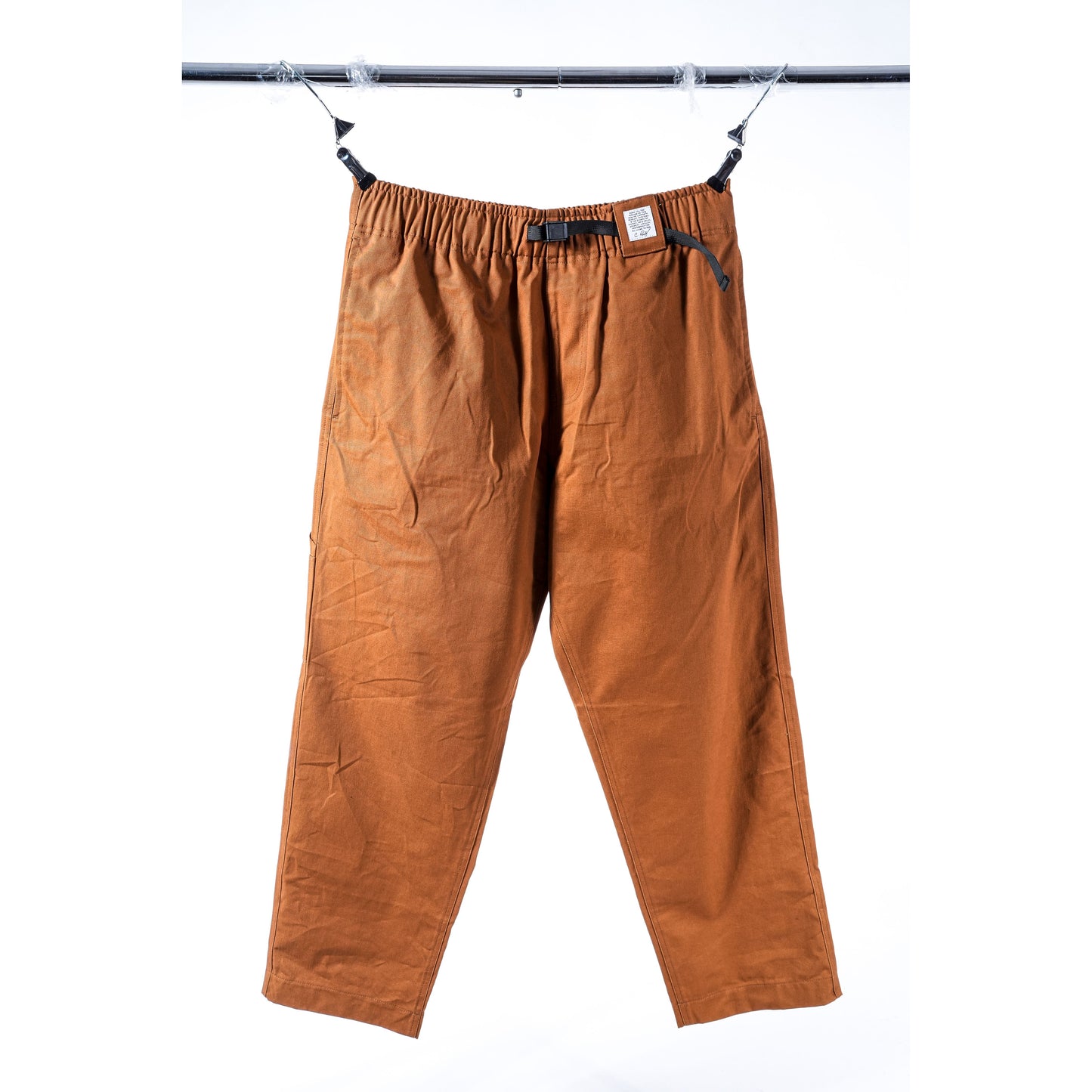 A pair of loose-fitting, beige baggy skate pants with an elastic waistband and black drawstrings, hanging from a black metal rod against a white background.