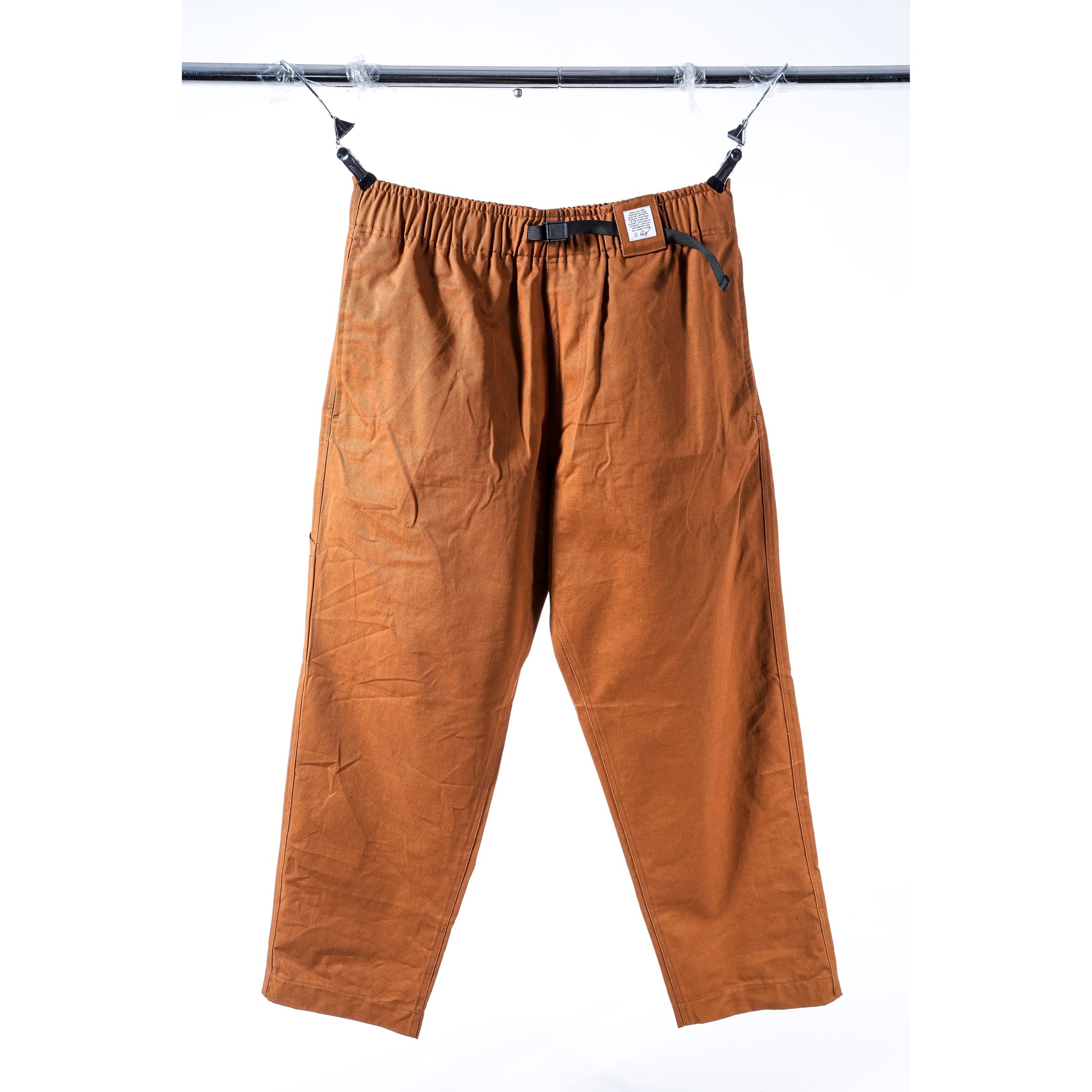 A pair of loose-fitting, beige baggy skate pants with an elastic waistband and black drawstrings, hanging from a black metal rod against a white background.