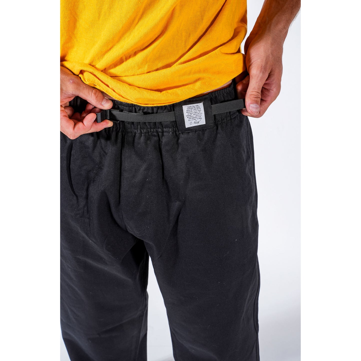 Close-up of a person wearing black baggy skate pants and a yellow shirt, adjusting the waistband with both hands. A small white rectangular label is visible at the front center of the waistband.