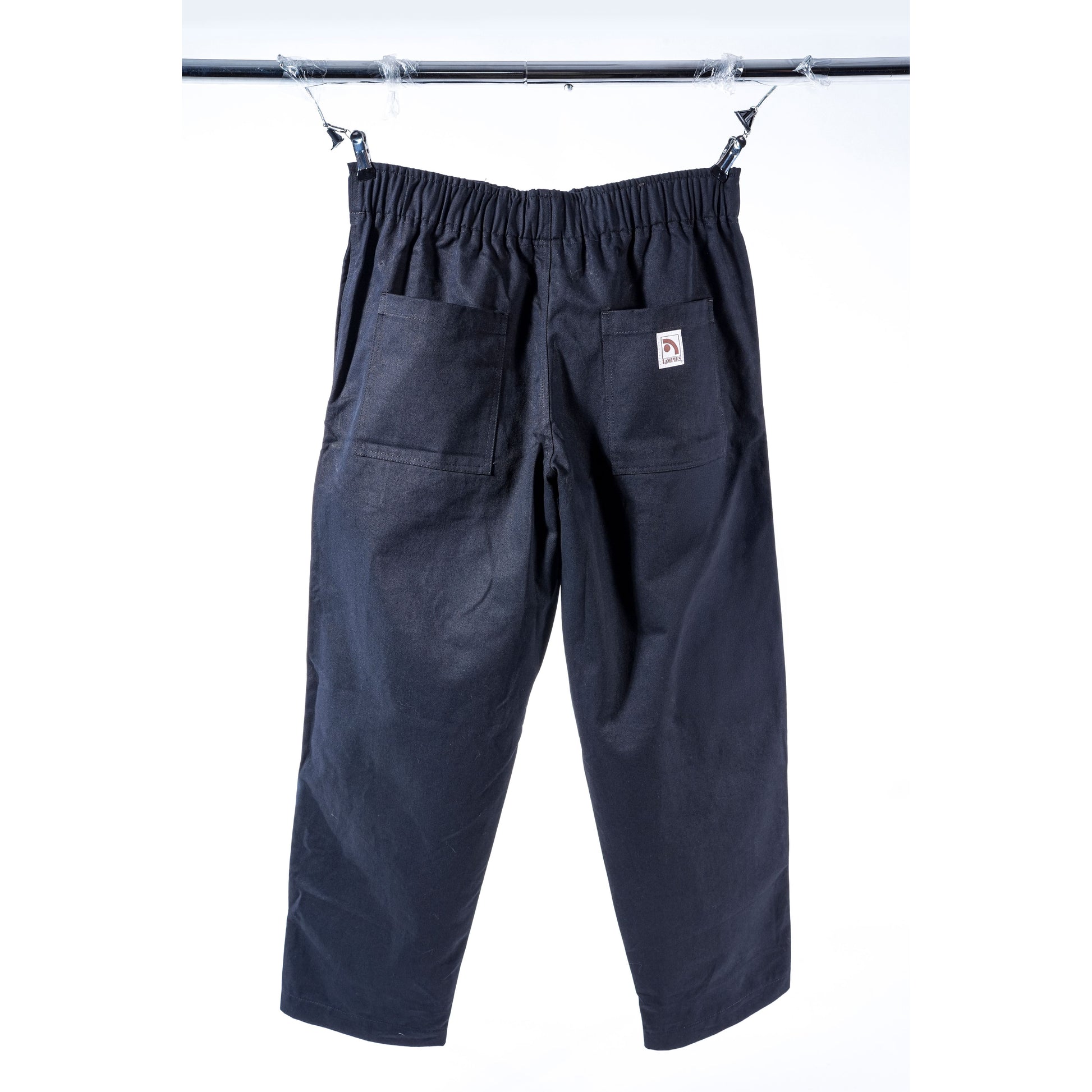 Black baggy skate pants with an elastic waistband, two large patch pockets, and a small white brand tag on the back pocket, hanging from clips on a metal bar against a white background.