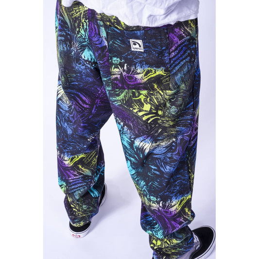 A close-up of a person wearing Limpies baggy skate pants with a vibrant, multicolor abstract print in purple, blue, green, and black. The photo is cropped from the waist down, showing the pants paired with a white t-shirt and black sneakers.