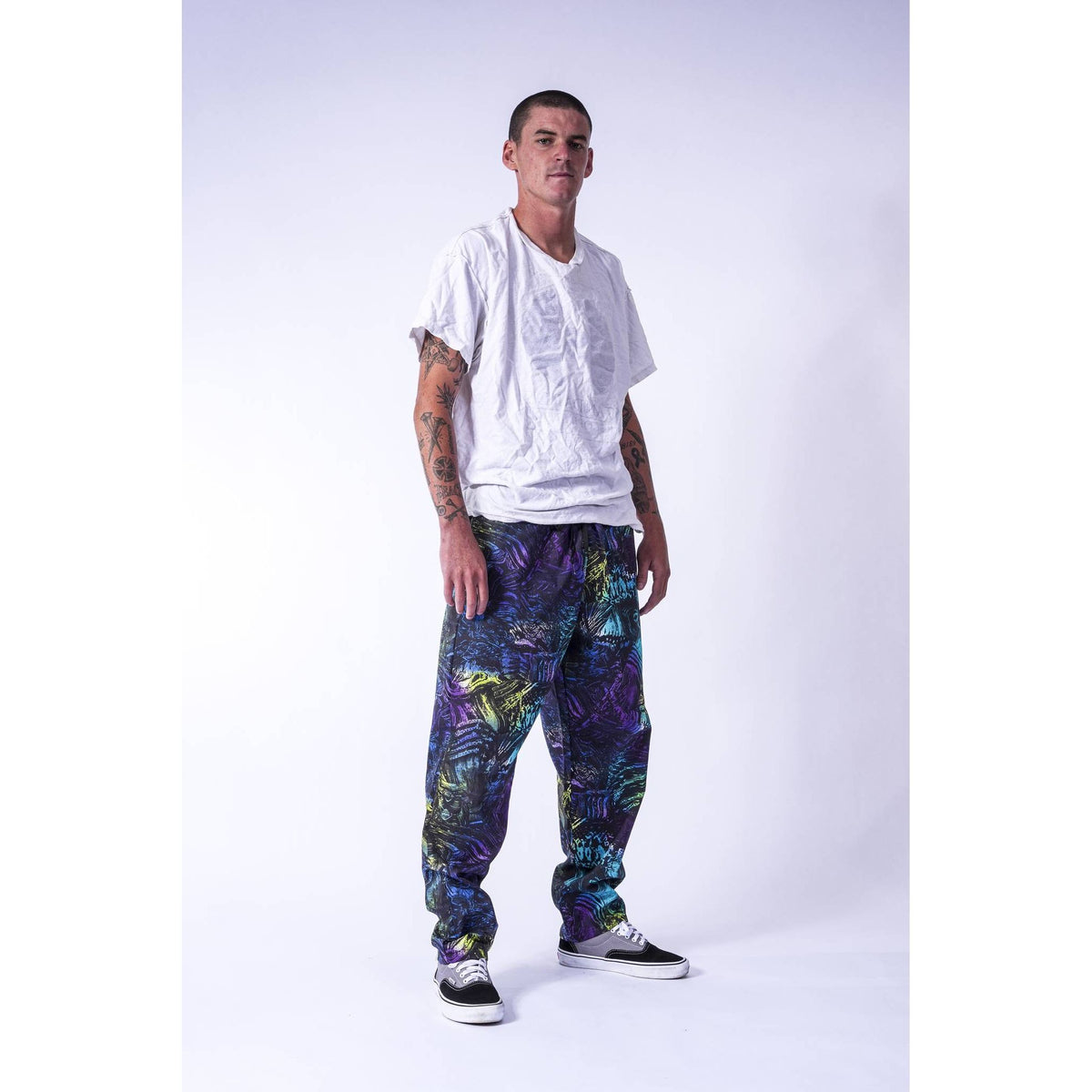 A man modeling a white t-shirt and colorful abstract print Limpies baggy skate pants with black and white sneakers, standing against a light gray studio background.