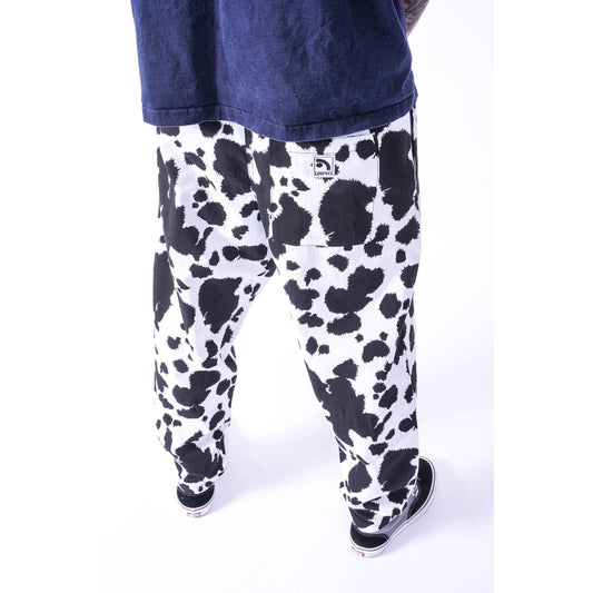 A person shown from the back wearing loose black-and-white cow-print  Limpies baggy skate pants, a dark navy T-shirt, and black sneakers against a clean white studio background.