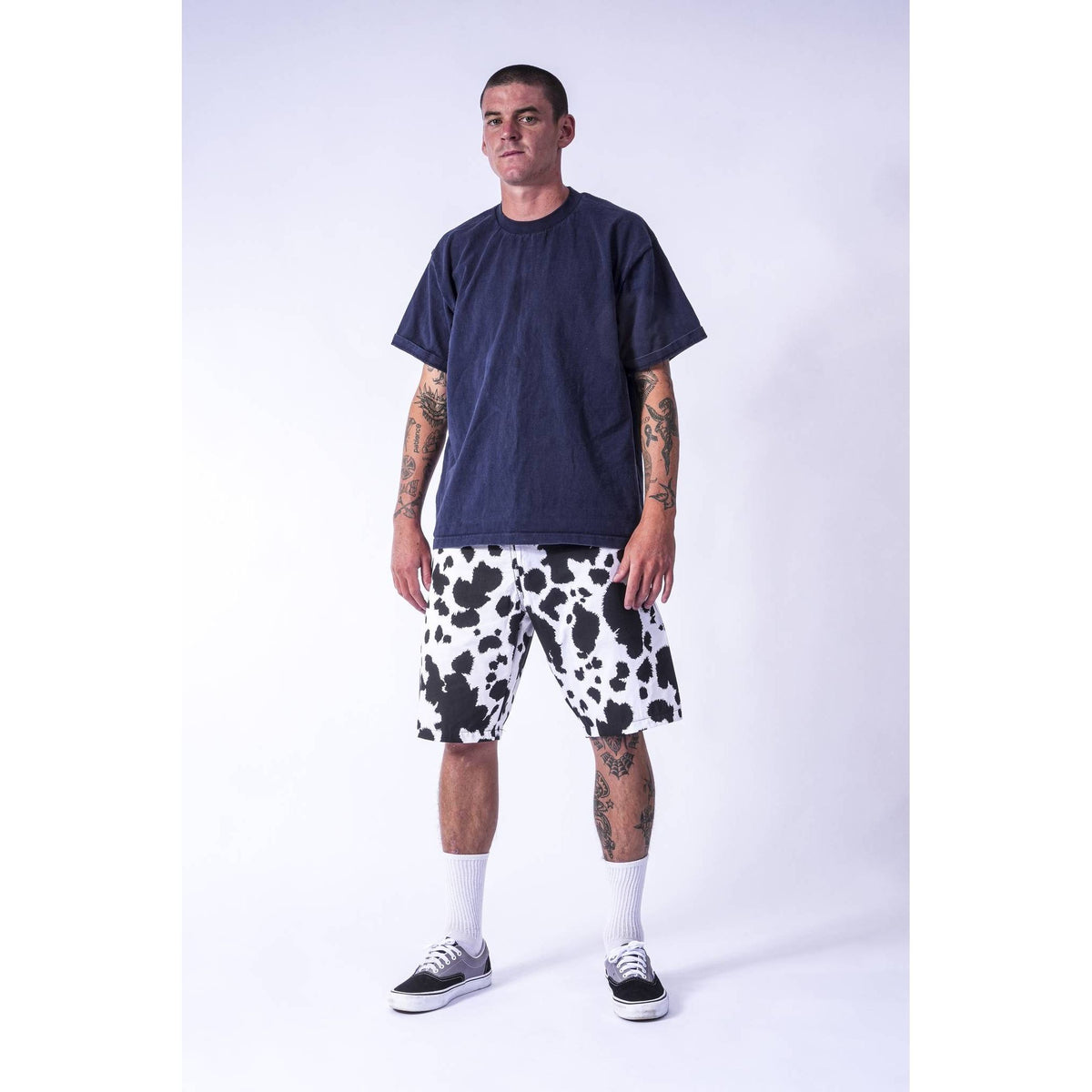 A man modeling a dark navy blue t-shirt and black and white cow-print baggy  Limpies skate shorts against a light gray studio background.