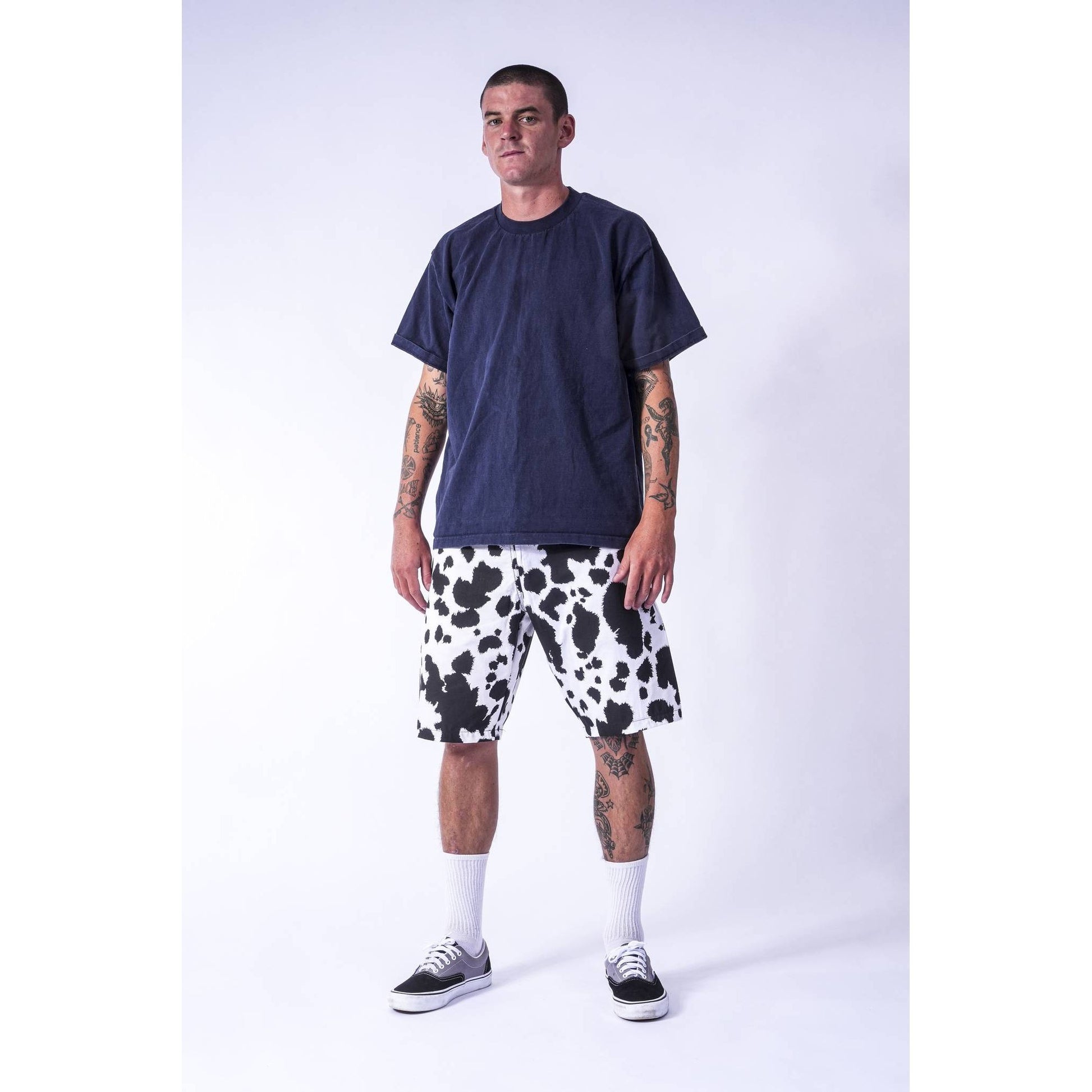 A man modeling a dark navy blue t-shirt and black and white cow-print baggy  Limpies skate shorts against a light gray studio background.
