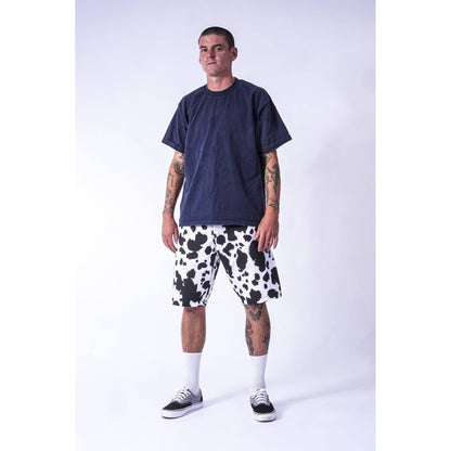 A man modeling a dark navy blue t-shirt and black and white cow-print baggy  Limpies skate shorts against a light gray studio background.