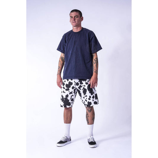 A man modeling a dark navy blue t-shirt and black and white cow-print baggy  Limpies skate shorts against a light gray studio background.