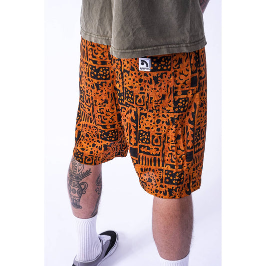 A person wearing orange and black patterned Limpies baggy skate shorts, an olive-green t-shirt, white socks, and black sneakers, shown from the waist down against a white studio background.