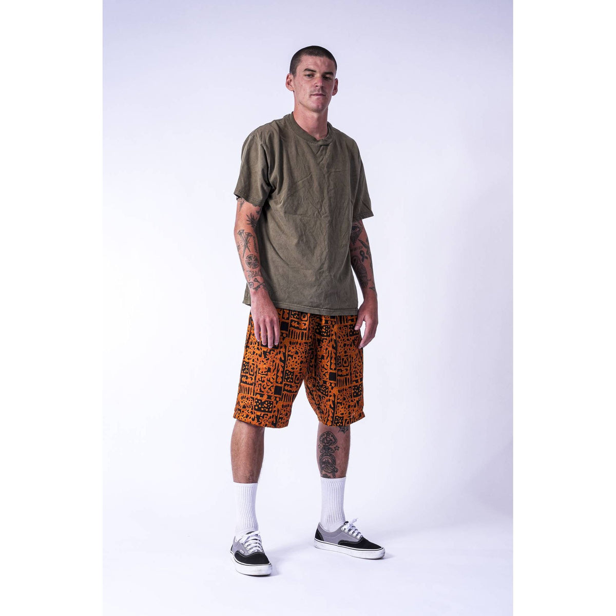 A man modeling a loose-fit olive-green t-shirt and orange shorts with a black graffiti-style pattern Limpies baggy skate shorts, paired with white socks and black-and-white sneakers, against a light gray studio background.