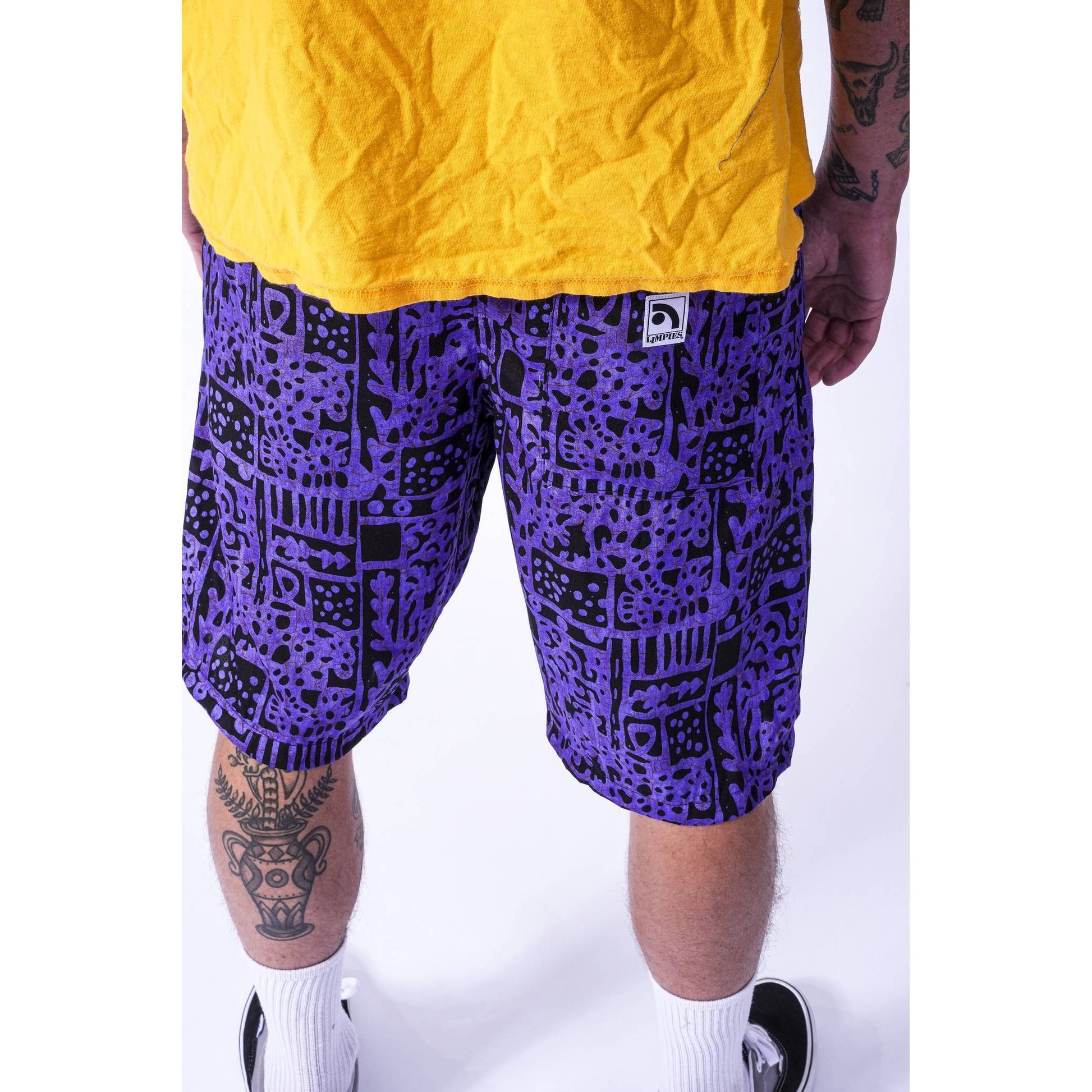 A person wearing purple and black patterned Limpies baggy skate shorts with a yellow t-shirt and white socks, shown from the waist down against a white studio background.