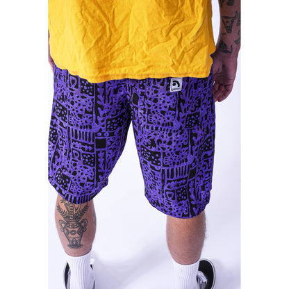 A person wearing purple and black patterned Limpies baggy skate shorts with a yellow t-shirt and white socks, shown from the waist down against a white studio background.