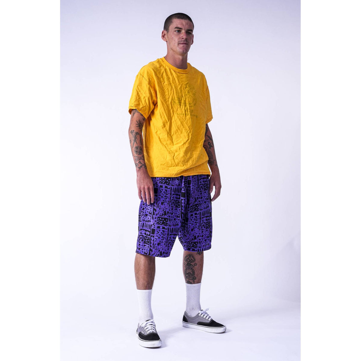 A street skater man modeling a bright yellow t-shirt and purple patterned Limpies baggy skate shorts against a light gray studio background.