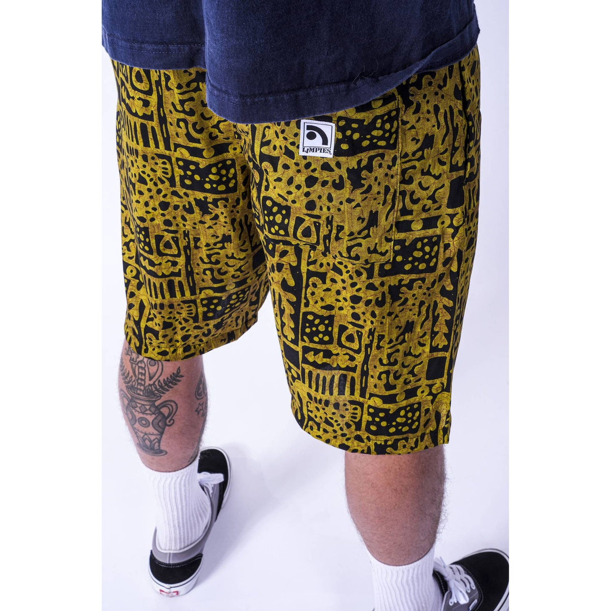 A person wearing yellow and black patterned  Limpies baggy skate shorts, a navy t-shirt, white socks, and black sneakers, shown from the waist down against a white studio background. The shorts feature a small white square logo patch on the left leg with the brand name 'Limpies'.