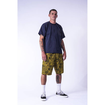 A street skater man modeling a dark navy blue t-shirt and yellow Limpies baggy skate shorts with a black graffiti-style pattern, paired with white socks and black sneakers, against a plain white studio background.