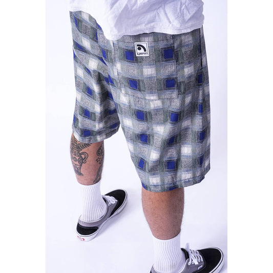 A person wearing gray and blue plaid Limpies baggy skate shorts with a white logo patch on the left leg, styled with a white t-shirt, white socks, and black sneakers against a white studio background.