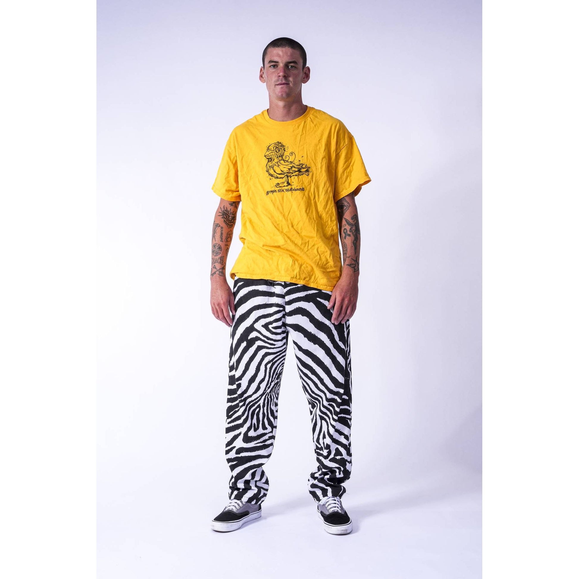 A man modeling a bright yellow graphic t-shirt and black and white zebra-print baggy skate pants against a light gray studio background.
