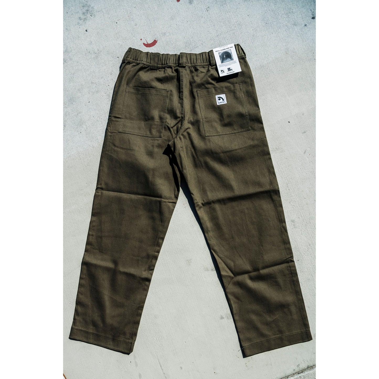 olive Green baggy skate pants with a brand tag on a gray concrete surface
