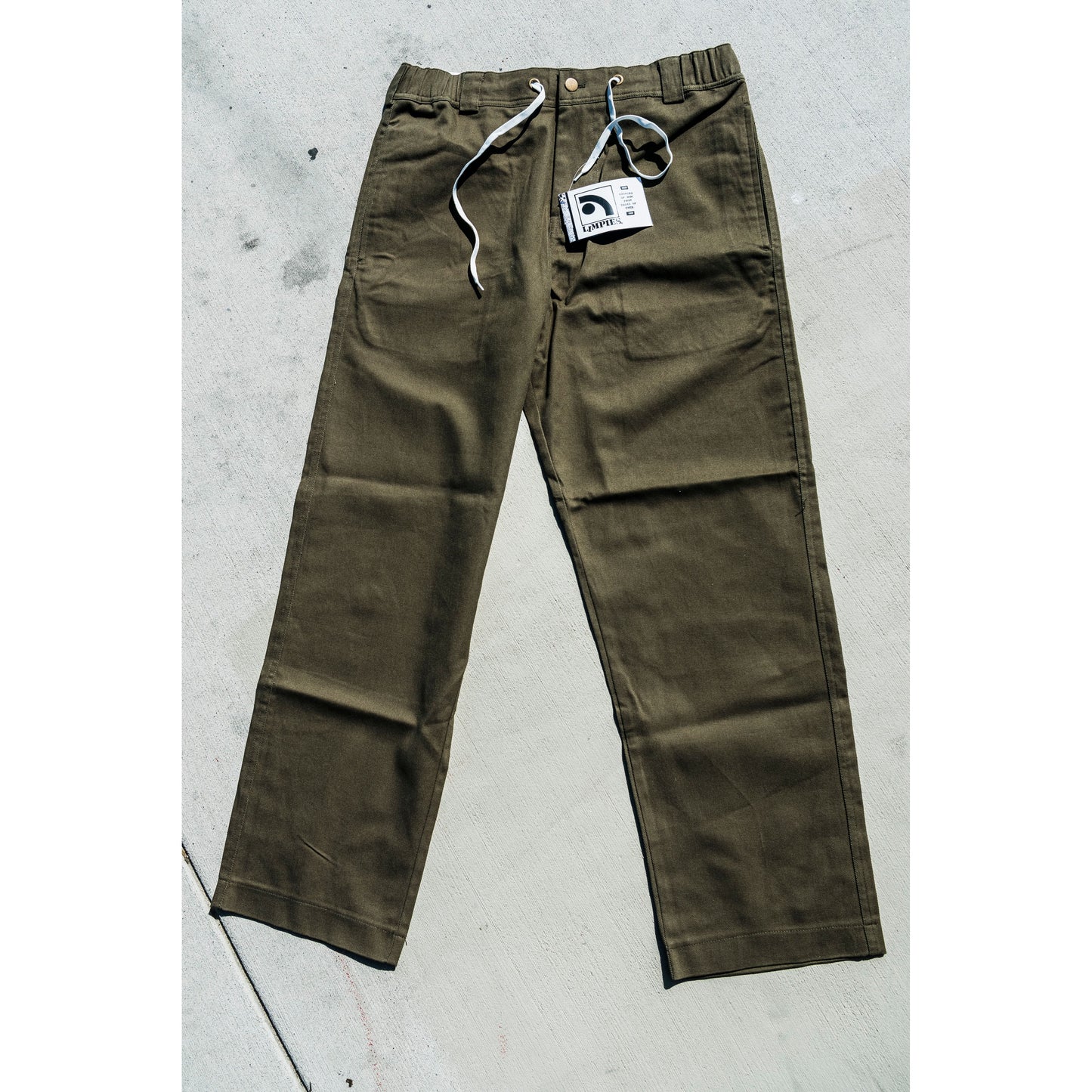 olive Green baggy skate pants with a tag on a gray concrete surface