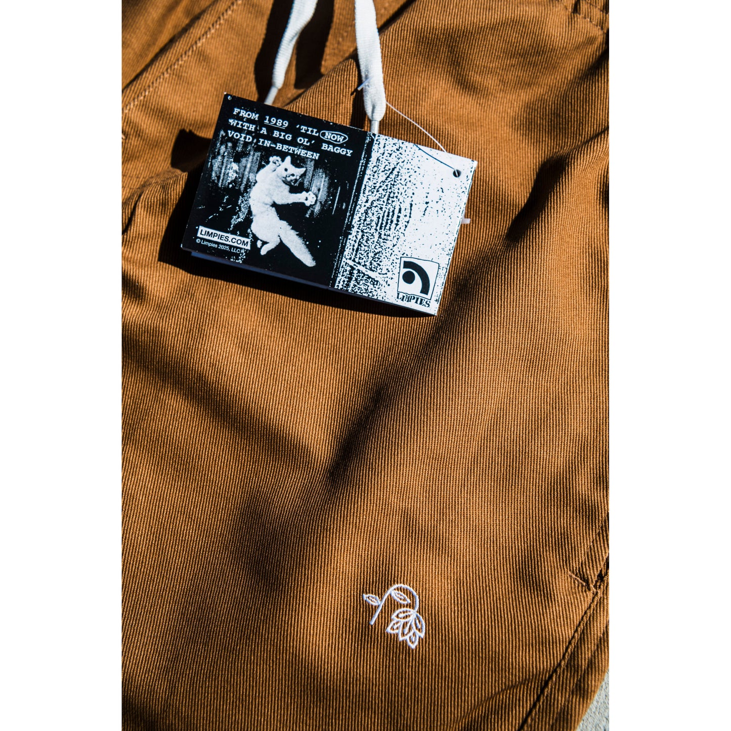 Close-up of a brown baggy skate pants with a flower embroidery and with a brand label featuring a flying cat graphic and text.