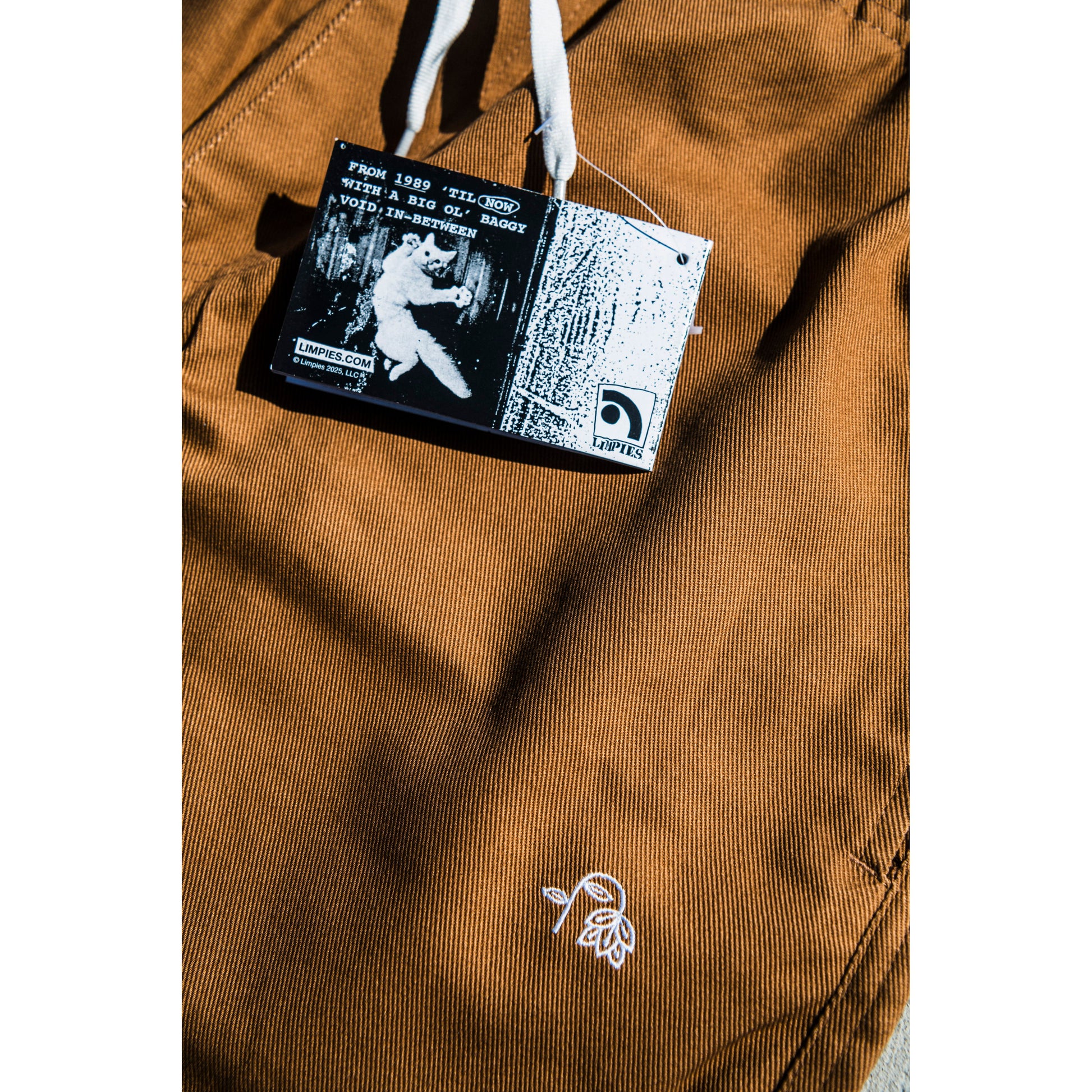 Close-up of a brown baggy skate pants with a flower embroidery and with a brand label featuring a flying cat graphic and text.