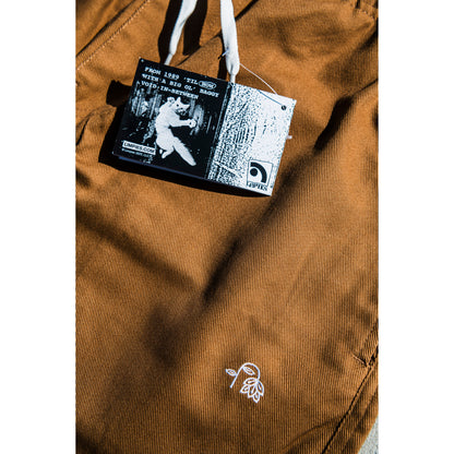 Close-up of a brown baggy skate pants with a flower embroidery and with a brand label featuring a flying cat graphic and text.
